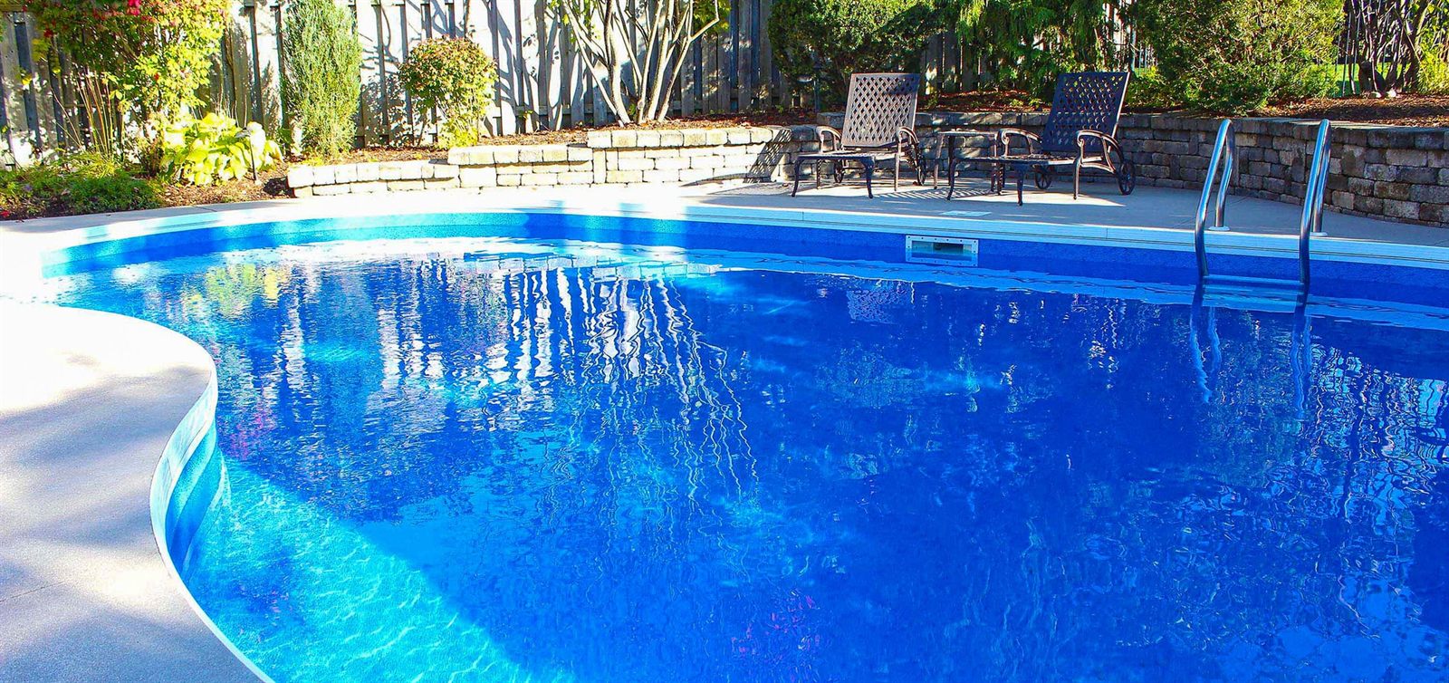 An in-ground pool finished with a bright blue Merlin vinyl liner, with a paved patio and retaining wall behind it